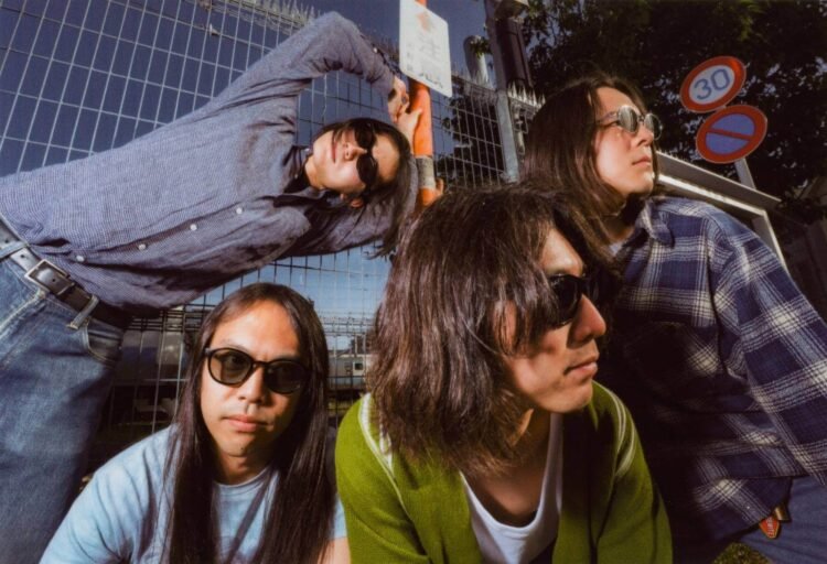 Band photo of the four members of DYGL posing in front of a camera, but most are not looking into the lens and two of them are even looking away. The musicians are dressed casually and all wearing sun glasses. They are posing in front of a gate and a train is visible in the background. Photography: Earl Standerford