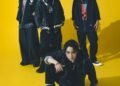 Group photo of the band SPYAIR, all dressed in dark coloured clothing against a yellow background. From left to right: KENTA (Drums), MOMIKEN (Bass player), YOSUKE (Vocalist), UZ (Guitarist & Programming). Whilst vocalist YOSUKE is sitting on the floor, the other band members are standing close together behind him. © Sony Music
