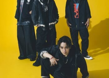 Group photo of the band SPYAIR, all dressed in dark coloured clothing. From left to right: KENTA (Drums), MOMIKEN (Bass player), YOSUKE (Vocalist), UZ (Guitarist & Programming). Whilst vocalist YOSUKE is sitting on the floor, the other band members are standing close together behind him.