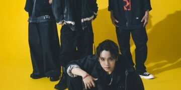 Group photo of the band SPYAIR, all dressed in dark coloured clothing. From left to right: KENTA (Drums), MOMIKEN (Bass player), YOSUKE (Vocalist), UZ (Guitarist & Programming). Whilst vocalist YOSUKE is sitting on the floor, the other band members are standing close together behind him.