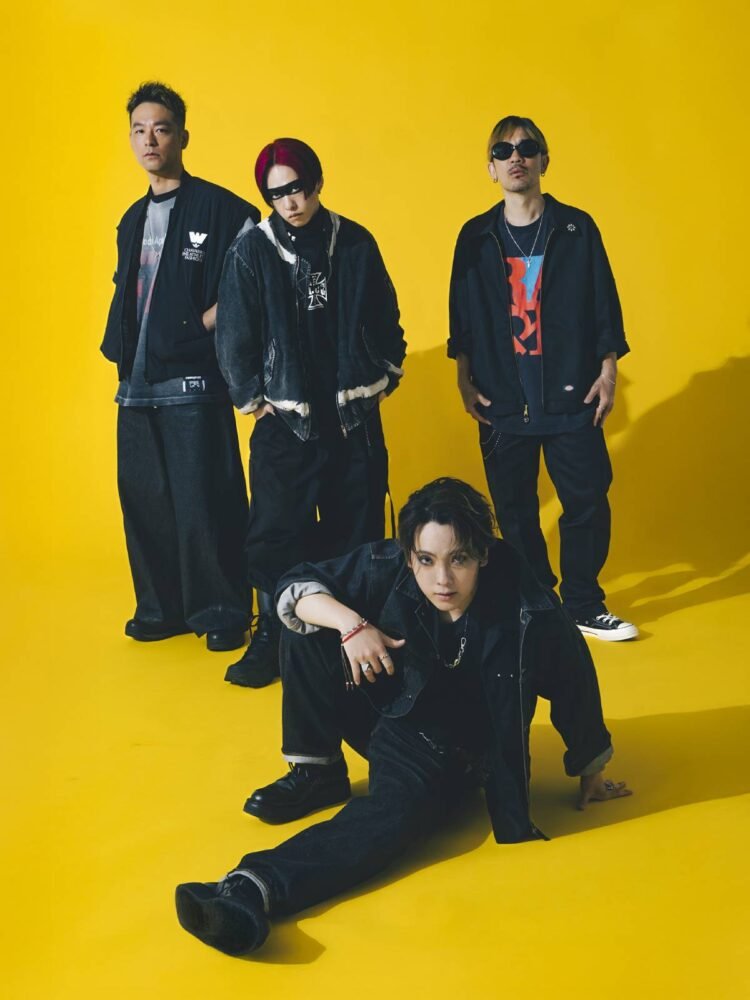 Group photo of the band SPYAIR, all dressed in dark coloured clothing against a yellow background. From left to right: KENTA (Drums), MOMIKEN (Bass player), YOSUKE (Vocalist), UZ (Guitarist & Programming). Whilst vocalist YOSUKE is sitting on the floor, the other band members are standing close together behind him. © Sony Music
