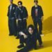 Group photo of the band SPYAIR, all dressed in dark coloured clothing. From left to right: KENTA (Drums), MOMIKEN (Bass player), YOSUKE (Vocalist), UZ (Guitarist & Programming). Whilst vocalist YOSUKE is sitting on the floor, the other band members are standing close together behind him.