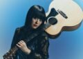 Singer and guitarist Upiko poses with her acoustic guitar. The guitar is resting against her left shoulder whilst she holds the neck of the guitar. She is dressed in black and blue, with elements of lace and wat seems to be velvet.