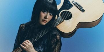 Singer and guitarist Upiko poses with her acoustic guitar. The guitar is resting against her left shoulder whilst she holds the neck of the guitar. She is dressed in black and blue, with elements of lace and wat seems to be velvet.