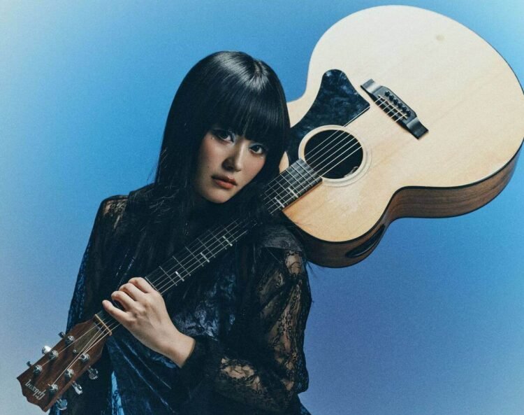 Singer and guitarist Upiko poses with her acoustic guitar. The guitar is resting against her left shoulder whilst she holds the neck of the guitar. She is dressed in black and blue, with elements of lace and wat seems to be velvet.