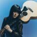 Singer and guitarist Upiko poses with her acoustic guitar. The guitar is resting against her left shoulder whilst she holds the neck of the guitar. She is dressed in black and blue, with elements of lace and wat seems to be velvet.