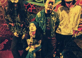 The photo shows the four members of Hibushibire posing for the camera, three of them are standing, while one of them, the drummer, is sitting on a stool. The photo is colourful, because of the background. The wall is decorated with paint and graffiti and seems to be a live bar or venue.