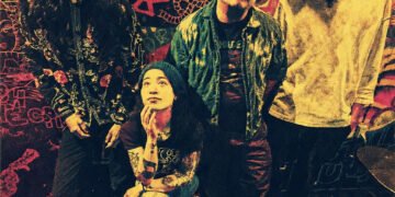 The photo shows the four members of Hibushibire posing for the camera, three of them are standing, while one of them, the drummer, is sitting on a stool. The photo is colourful, because of the background. The wall is decorated with paint and graffiti and seems to be a live bar or venue.