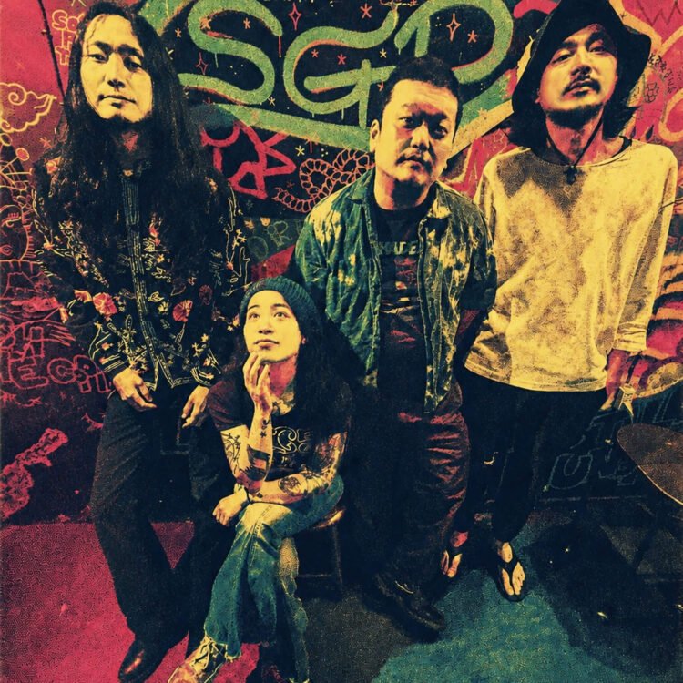 The photo shows the four members of Hibushibire posing for the camera, three of them are standing, while one of them, the drummer, is sitting on a stool. The photo is colourful, because of the background. The wall is decorated with paint and graffiti and seems to be a live bar or venue.