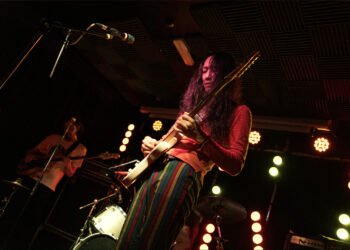 Bandleader of Hibushibire, Changchang, plays the guitar on the stage of The Victoria in Birmingham. The musician is wearing colourful clothing, a reddish shirt and vertical striped pants in the colours green, red, and yellow. In the background there is another member, Tetsuji Toyoda, playing the bass. Photography JJ Grant (wonderlens)