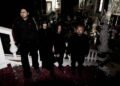 The four members of kokeshi standing on a staircase of an old building that is filled with statues, while it seems to be derelict and maybe also declared unfit for habitation. All members are dressed in black clothing and not looking into the camera lens. © kokeshi