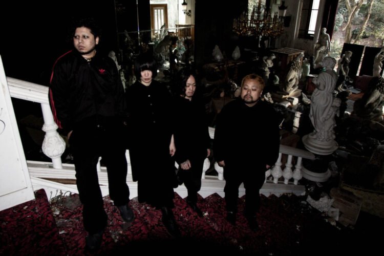 The four members of kokeshi standing on a staircase of an old building that is filled with statues, while it seems to be derelict and maybe also declared unfit for habitation. All members are dressed in black clothing and not looking into the camera lens. © kokeshi