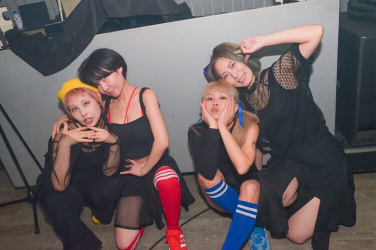 The four members of cheer punk band Ni-hao!!!! posing together before or after a show. They are all dressed in black clothing and wear coloured socks.  © Photo by Yukiko Ono