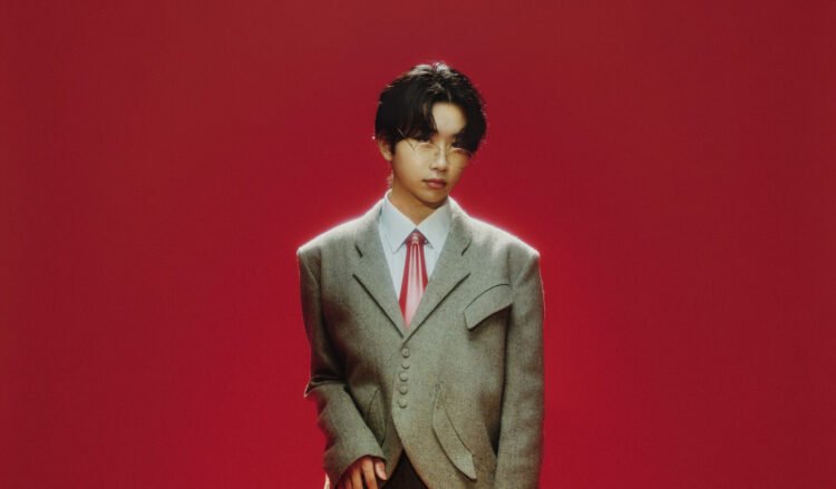 Singer-songwriter AKASAKI is wearing a greyish tweed blazer with a red glossy necktie and glasses. He is posing in front of a red background.