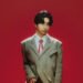 Singer-songwriter AKASAKI is wearing a greyish tweed blazer with a red glossy necktie and glasses. He is posing in front of a red background.