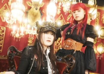 Photo of two members of FATE GEAR, the bandleader Captain Mina (sitting on a chair) and red-headed vocalist Hime Hashimura standing behind the chair. The musicians are dressed in steampunk-style outfits and posing in a room that is filled with chandeliers and gold-coloured decorations against a red background. © Steam Steel Records