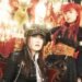 Photo of two members of FATE GEAR, the bandleader Captain Mina (sitting on a chair) and red-headed vocalist Hime Hashimura standing behind the chair. The musicians are dressed in steampunk-style outfits and posing in a room that is filled with chandeliers and gold-coloured decorations against a red background. © Steam Steel Records