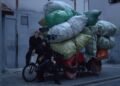 The three members of She Her Her Hers are posing with a heavily loaded bicycle or tricycle in a quiet alley. One of the members seems to be sitting on the saddle, while the other member is sitting on the steering wheel. The third band member is crouching next to the bike. All members are dressed in dark clothing, while the heavy load consist of colourful bags making it an interesting contrast. © She Her Her Hers