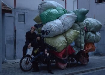 The three members of She Her Her Hers posing with a heavily loaded bicycle or tricycle in a quiet alley. One of the members seems to be sitting on the saddle, while the other member is sitting on the steering wheel. The third band member is crouching next to the bike. All members are dressed in dark clothing, while the heavy load consist of colourful bags making it an interesting contrast. © She Her Her Hers