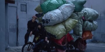 The three members of She Her Her Hers posing with a heavily loaded bicycle or tricycle in a quiet alley. One of the members seems to be sitting on the saddle, while the other member is sitting on the steering wheel. The third band member is crouching next to the bike. All members are dressed in dark clothing, while the heavy load consist of colourful bags making it an interesting contrast. © She Her Her Hers
