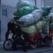 The three members of She Her Her Hers posing with a heavily loaded bicycle or tricycle in a quiet alley. One of the members seems to be sitting on the saddle, while the other member is sitting on the steering wheel. The third band member is crouching next to the bike. All members are dressed in dark clothing, while the heavy load consist of colourful bags making it an interesting contrast. © She Her Her Hers