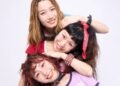The photo features the three women of THE LET'S GO's posing very closely together. They are standing one behind the other, but are sitting or standing at different heights and leaning against one another, so that their faces are clearly visible and arranged in a row. They are all dressed stylishly and colourful against a white wall. Photography by 安藤信之 (Ando Nobuyuki), makeup by monmo.