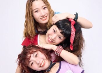 The photo features the three women of THE LET'S GO's posing very closely together. They are standing one behind the other, but are sitting or standing at different heights and leaning against one another, so that their faces are clearly visible and arranged in a row. They are all dressed stylishly and colourful against a white wall. Photography by 安藤信之 (Ando Nobuyuki), makeup by monmo.