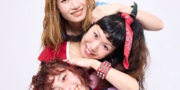 The photo features the three women of THE LET'S GO's posing very closely together. They are standing one behind the other, but are sitting or standing at different heights and leaning against one another, so that their faces are clearly visible and arranged in a row. They are all dressed stylishly and colourful against a white wall. Photography by 安藤信之 (Ando Nobuyuki), makeup by monmo.