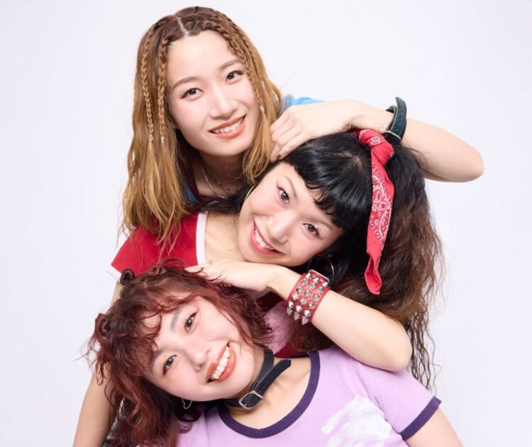 The photo features the three women of THE LET'S GO's posing very closely together. They are standing one behind the other, but are sitting or standing at different heights and leaning against one another, so that their faces are clearly visible and arranged in a row. They are all dressed stylishly and colourful against a white wall. Photography by 安藤信之 (Ando Nobuyuki), makeup by monmo.