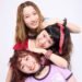 The photo features the three women of THE LET'S GO's posing very closely together. They are standing one behind the other, but are sitting or standing at different heights and leaning against one another, so that their faces are clearly visible and arranged in a row. They are all dressed stylishly and colourful against a white wall. Photography by 安藤信之 (Ando Nobuyuki), makeup by monmo.