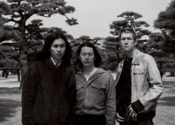 Black-and-white photo of the three members of Tō Yō facing the camera. They are standing together in a park with the background filled with trees. Photo by Takaumi Minagawa