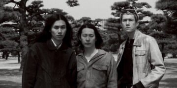 Black-and-white photo of the three members of Tō Yō facing the camera. They are standing together in a park with the background filled with trees. Photo by Takaumi Minagawa