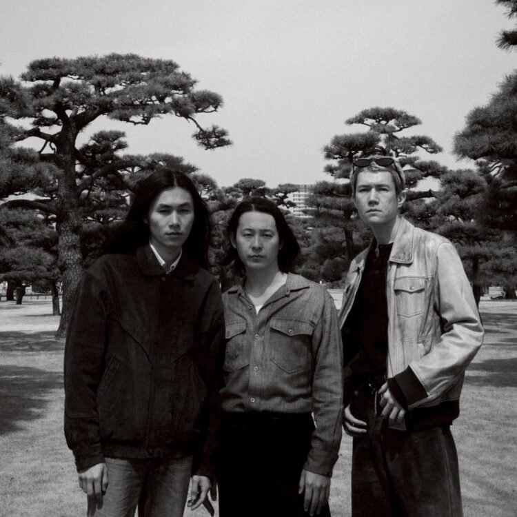 Black-and-white photo of the three members of Tō Yō facing the camera. They are standing together in a park with the background filled with trees. Photo by Takaumi Minagawa